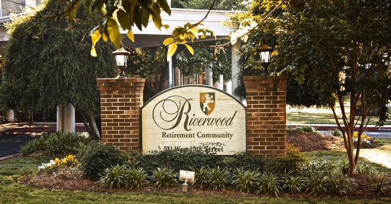 Entrance sign for Riverwood Retirement Community between brick pillars and lamps, surrounded by landscaping.