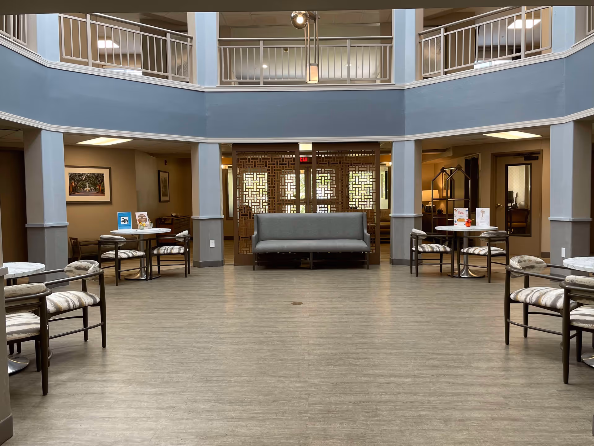 A spacious, well-lit common area in a senior living facility with a circular open ceiling above. The room features several small round tables with cushioned chairs arranged around the perimeter. In the center against the back wall is a gray cushioned bench with decorative wooden panels behind it. The walls are painted in neutral tones, and framed artwork is visible on the side walls. The floor is covered with light wood-style flooring.