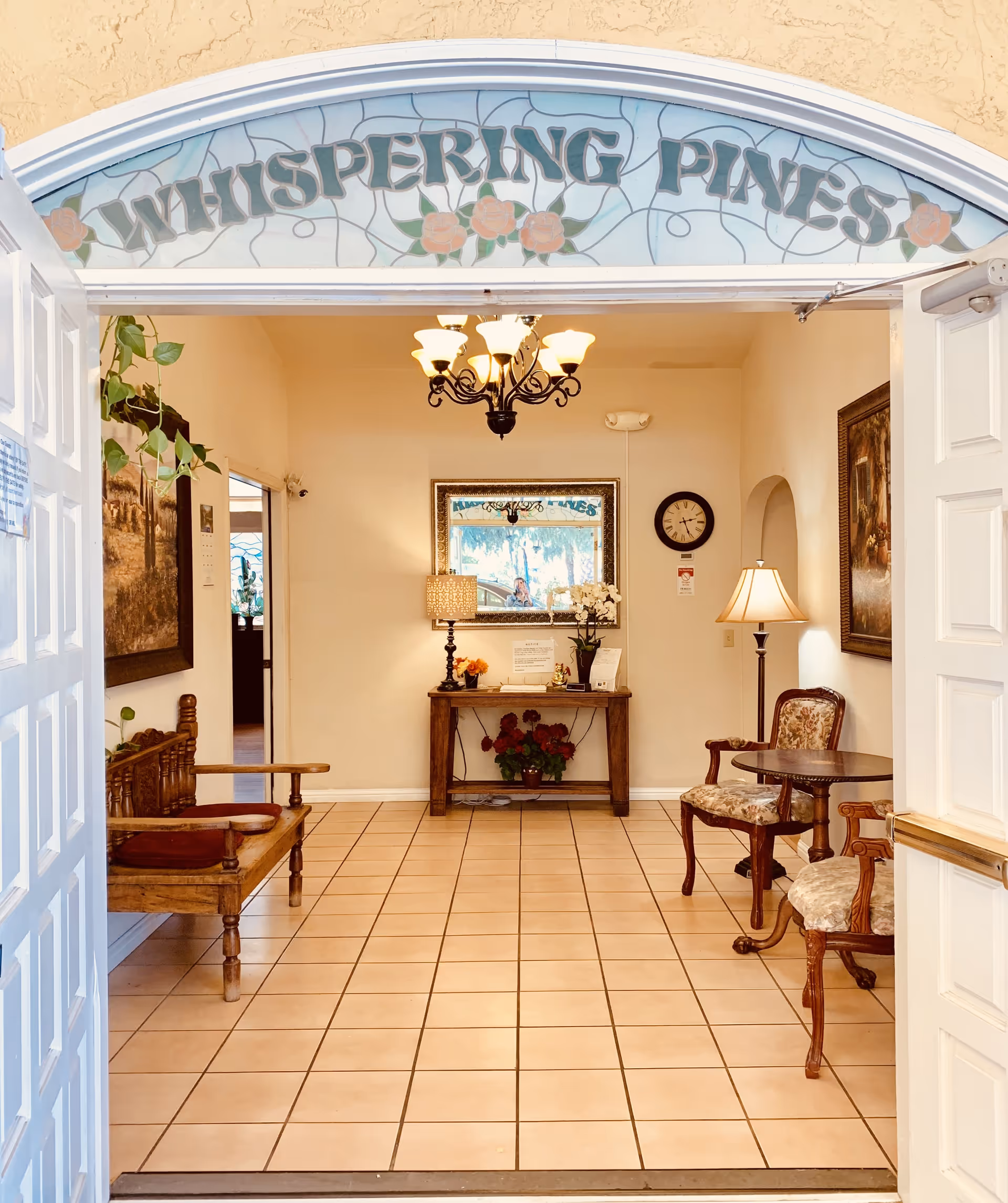 Entrance to Whispering Pines Inn showing an open double door leading into a tiled lobby area with wooden bench seating on the left, two upholstered chairs and a small round table on the right, a wooden console table with a lamp, flowers, and a mirror on the back wall, and a chandelier hanging from the ceiling.