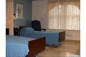 Two hospital-style beds with blue bedspreads in a shared resident room with a window, blinds, a chair, and a nightstand.
