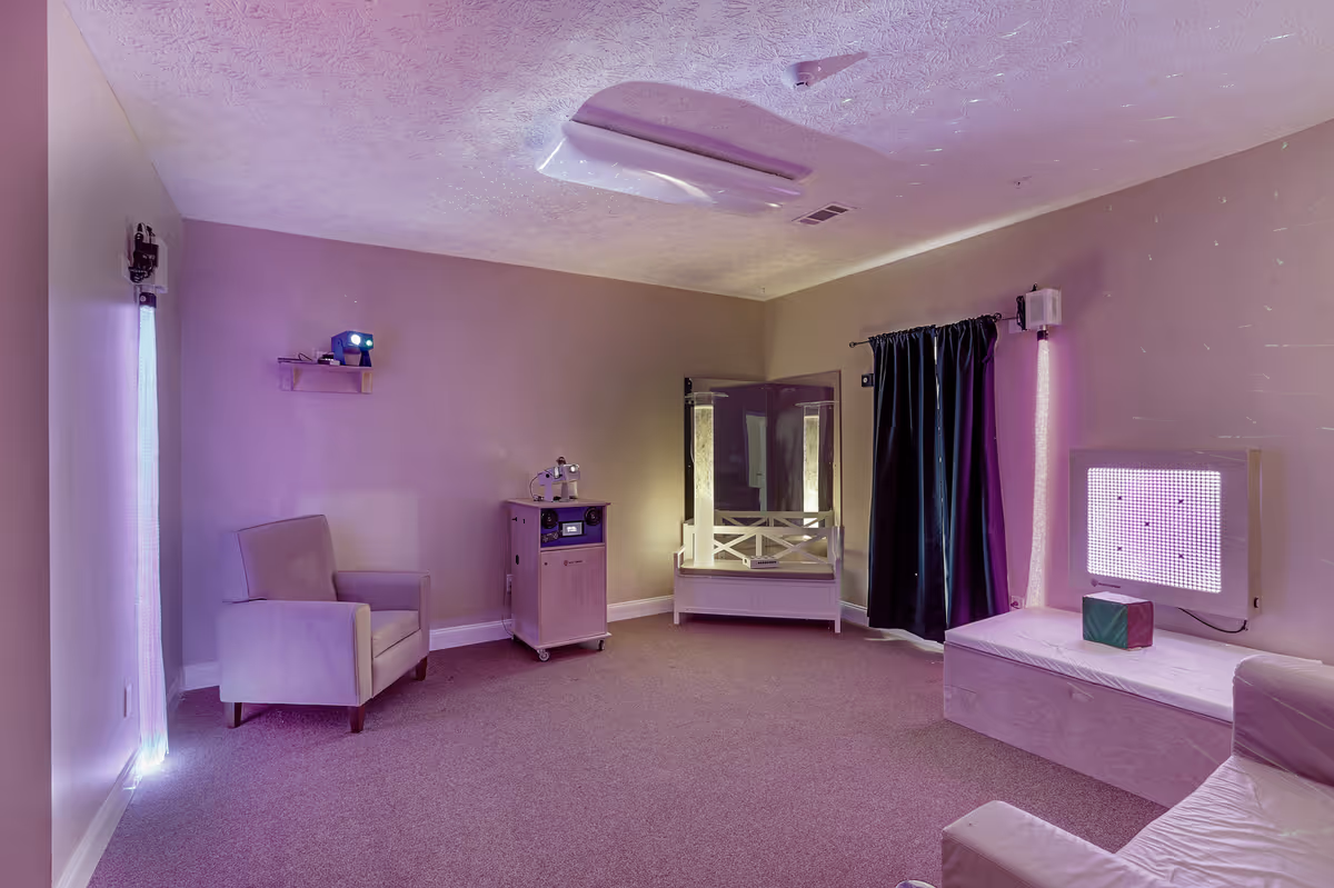A softly lit sensory room with purple lighting, featuring a beige armchair, a small wooden cabinet with electronic equipment, a mirrored corner with illuminated columns, a cushioned bench with a light panel emitting purple light, and a black curtain on the wall.