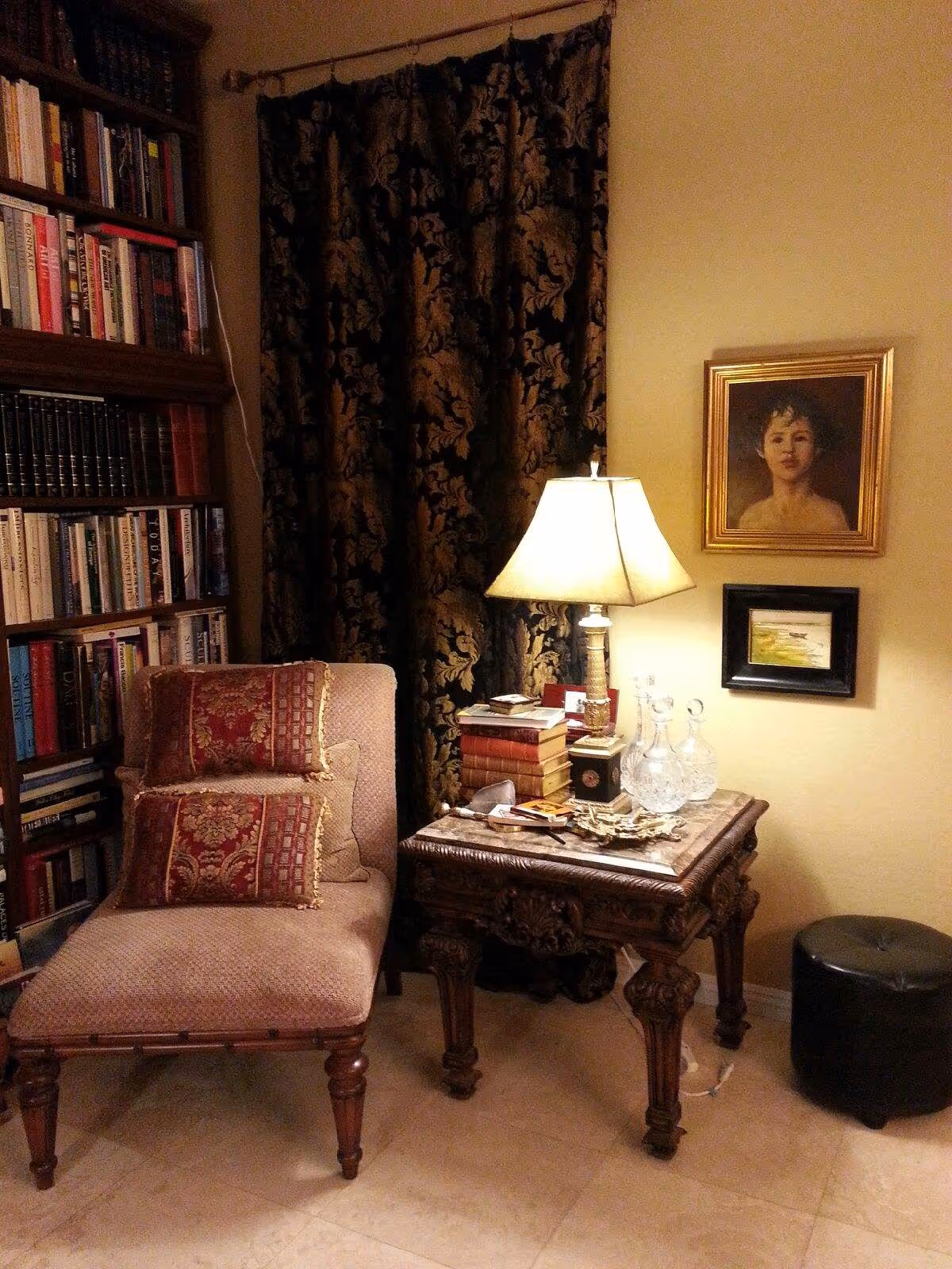 A cozy reading corner with a chaise lounge, bookshelf, ornate side table holding a lamp and glassware, and framed art on the wall.
