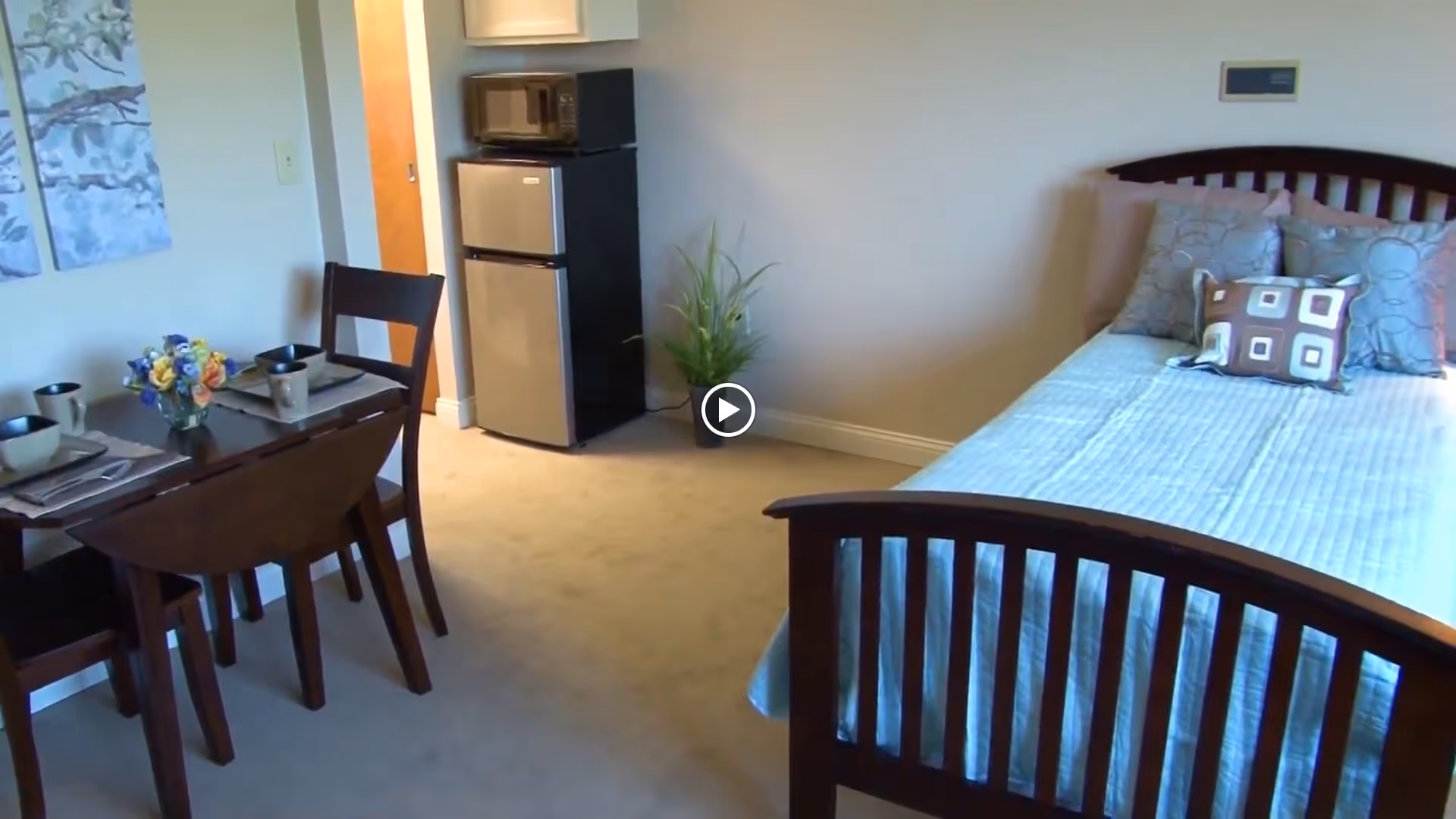 Studio-style senior living room with a wooden bed, small dining table set, and a mini fridge with microwave.