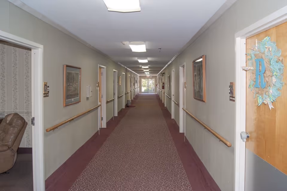 Long carpeted interior hallway with handrails, doors on both sides, framed pictures, and ceiling lights.