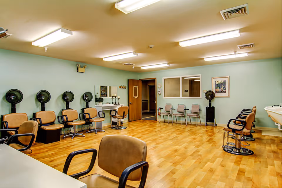 A spacious salon room with multiple hair-styling chairs, hooded dryers, mirrors, and waiting chairs on a wood floor.