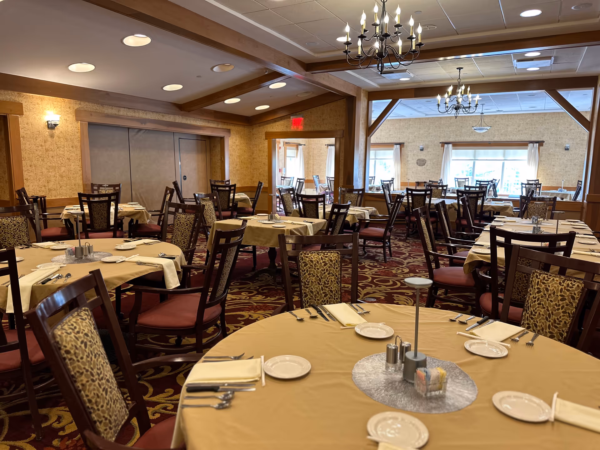 A spacious dining room with multiple round and rectangular tables covered with beige tablecloths. Each table is set with plates, napkins, silverware, and condiments. The chairs have wooden frames with patterned upholstery. The room features warm lighting, chandeliers, and large windows with white curtains allowing natural light to enter.