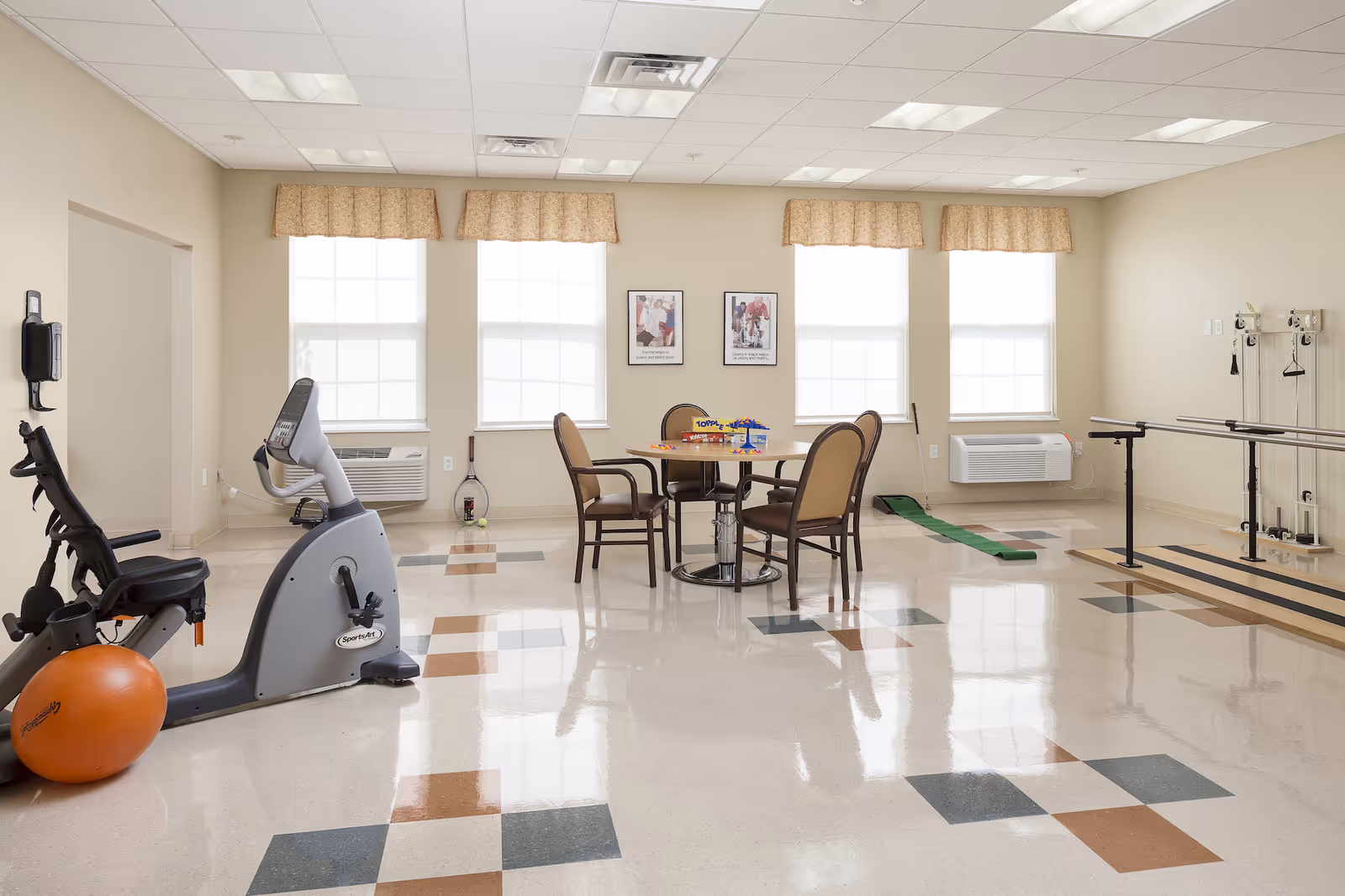 A bright room with exercise equipment including a recumbent bike and parallel bars, a round table with four chairs, and large windows with beige valances letting in natural light. The floor has a pattern of colored tiles, and there are two framed pictures on the wall between the windows.