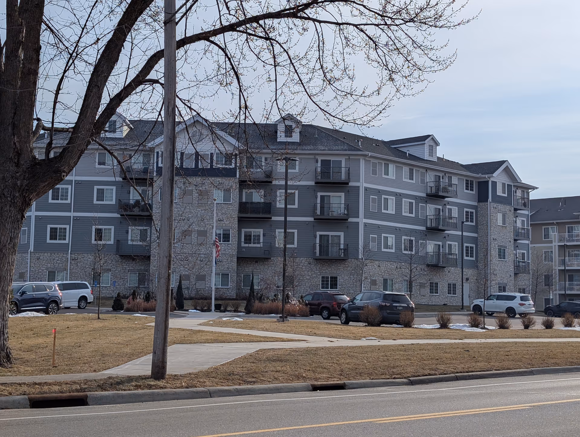 Exterior view of a multi-story senior housing building with gray and stone facade, balconies, and several parked cars in front. A large tree and a sidewalk are visible in the foreground under a cloudy sky.