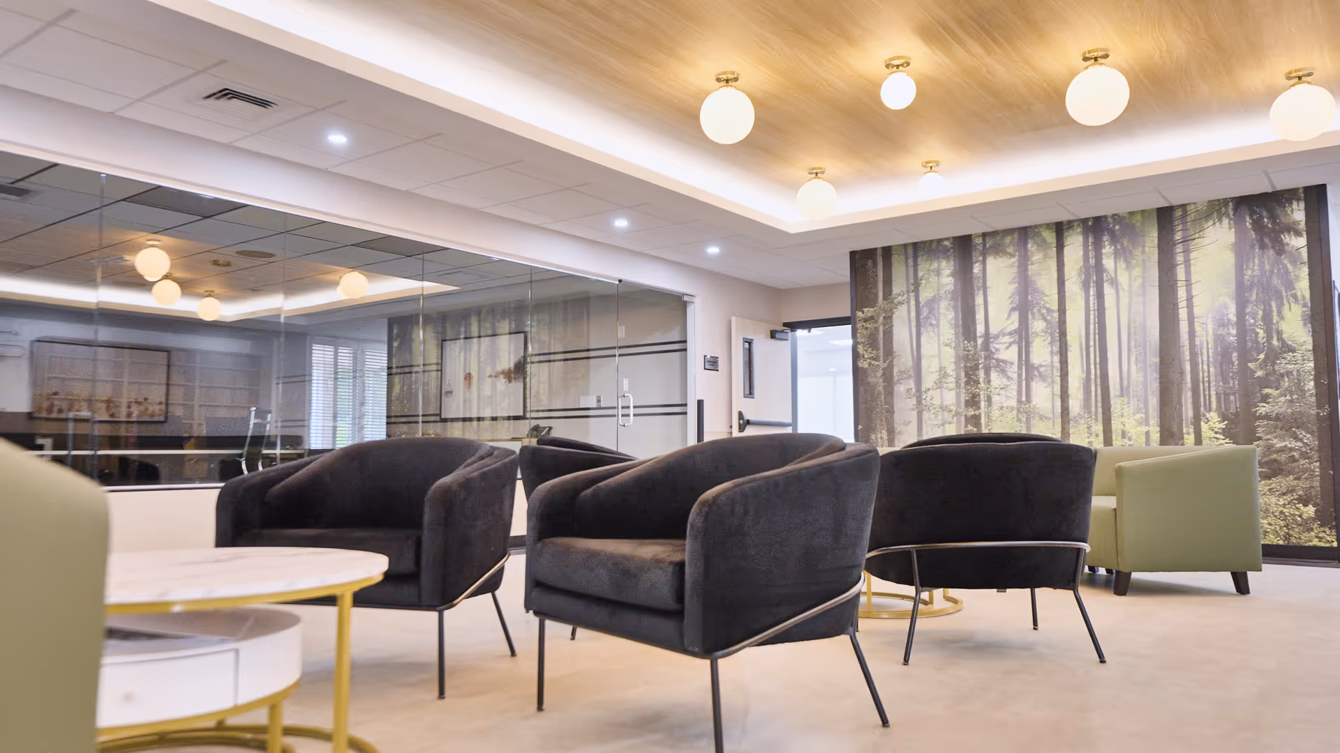A modern lounge area with black and green armchairs arranged around small round tables. The room features a large wall mural of a forest scene, a wooden ceiling with round pendant lights, and a mirrored wall reflecting the space.