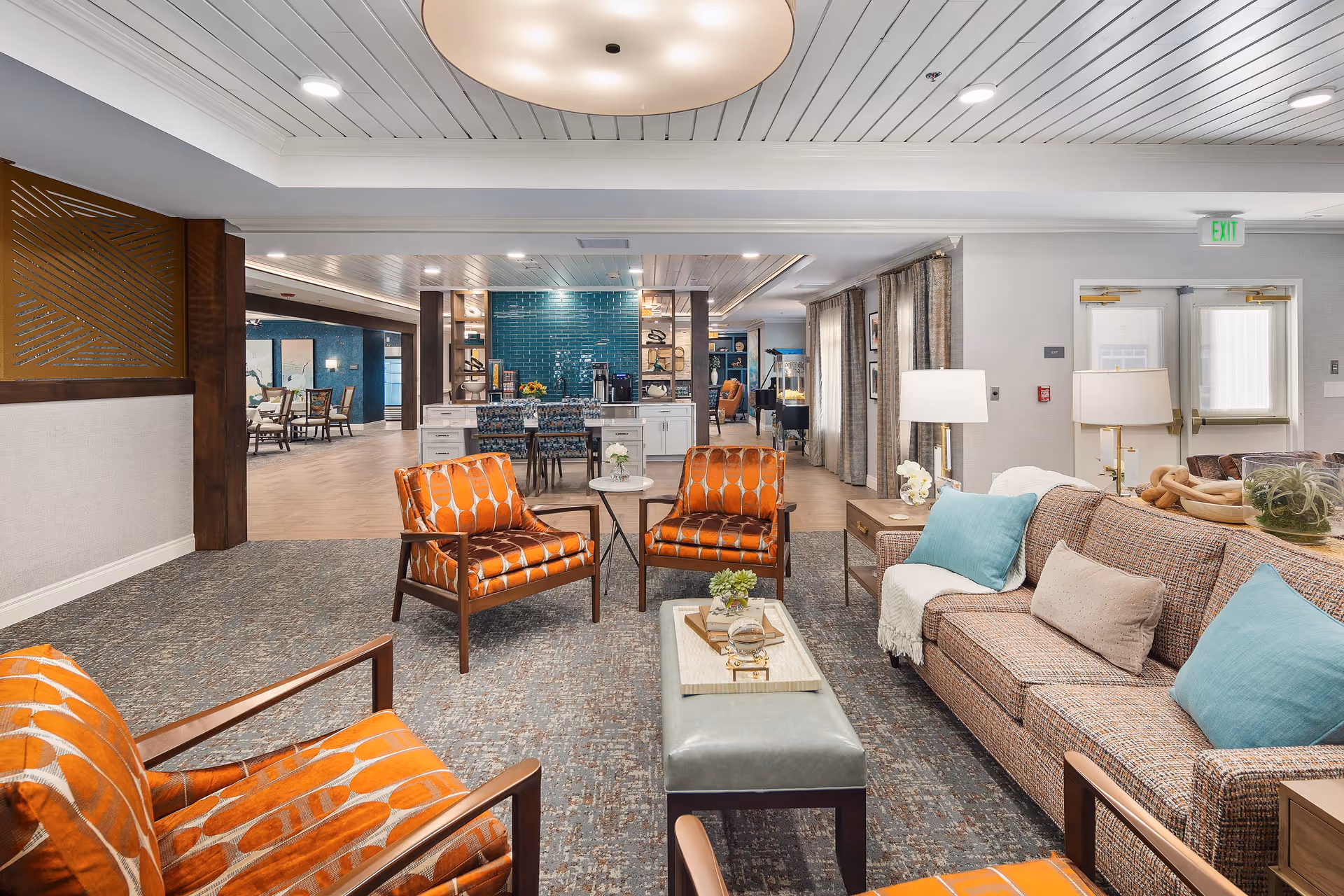 A spacious and well-lit senior living facility common area featuring a seating arrangement with a patterned sofa, blue and beige cushions, and three orange patterned armchairs around a small coffee table. In the background, there is a dining area with tables and chairs, a kitchen island with bar stools, and a teal tiled wall. The room has a modern ceiling with recessed lighting and large windows with curtains.