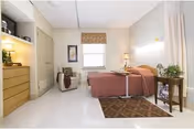 A neatly arranged bedroom in a nursing and rehab center featuring a single bed with a brown bedspread, a bedside table with a lamp and plant, a cushioned armchair with a blanket, a window with a valance, a dresser with shelves above it, and a patterned rug on the floor.
