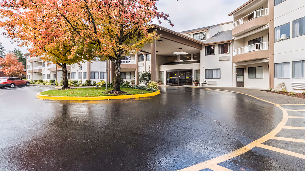 Covered front entrance of a multi-story retirement community with a circular driveway, wet pavement, and trees with autumn leaves.