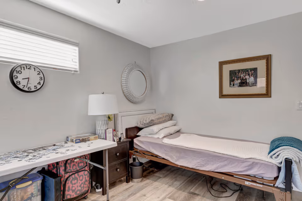 A small bedroom with a single bed against the wall, covered with a light purple sheet and a folded blanket at the foot. There are multiple pillows stacked at the head of the bed. Next to the bed is a dark wooden nightstand with a white lamp and some small items. Above the nightstand is a round decorative mirror on the wall. A framed family photo hangs on the adjacent wall. A folding table with a partially completed puzzle and puzzle box sits near the window, which has horizontal blinds. The floor is light wood, and the walls are painted light gray.