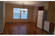 Interior view of a dining area and adjacent kitchen with wood floors, a chandelier, windows, refrigerator, and cabinets.