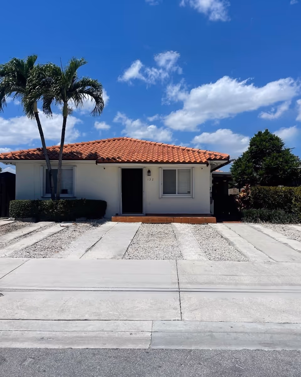Single-story house with a red tiled roof, white exterior walls, two windows with closed blinds, a black front door, and a small porch. There are two palm trees and some bushes in front of the house, with a clear blue sky and scattered clouds above. The driveway consists of concrete strips with gravel in between.