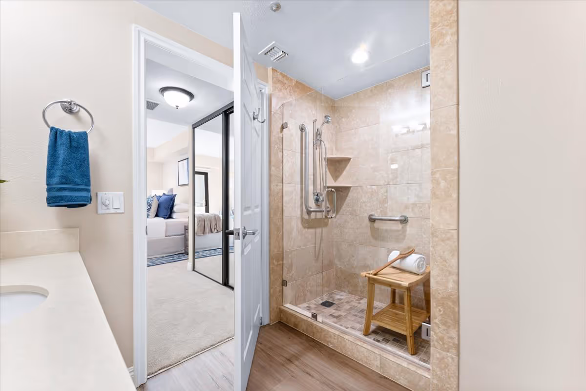 Tiled walk-in shower with grab bars and a wooden stool inside a bathroom, viewed through an open door into a bedroom.