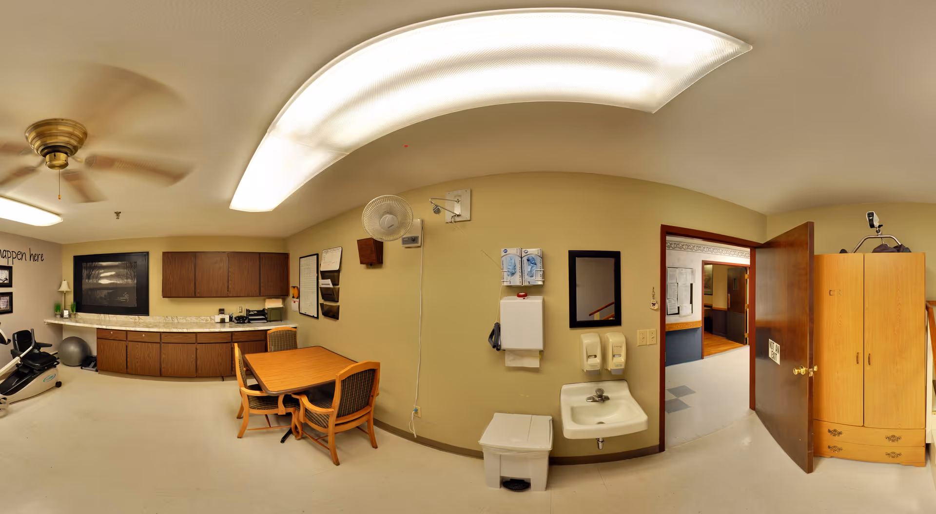 A room with exercise equipment including a stationary bike and an exercise ball, a wooden table with four chairs, a wall-mounted sink with soap dispensers and paper towel holder, cabinets along one wall, a ceiling fan, and an open door leading to a hallway.