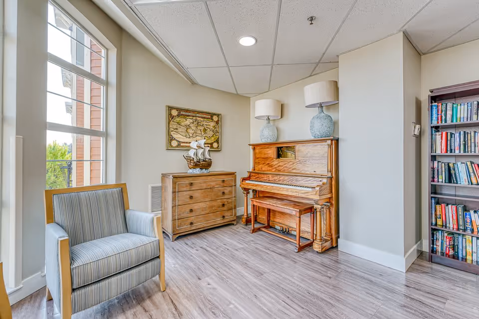 A cozy corner in a senior living facility featuring a wooden upright piano with a matching bench, two large table lamps with light-colored shades on top of the piano, a wooden chest of drawers with a decorative model ship, a framed vintage map on the wall, a striped upholstered armchair near a large window, and a bookshelf filled with books.