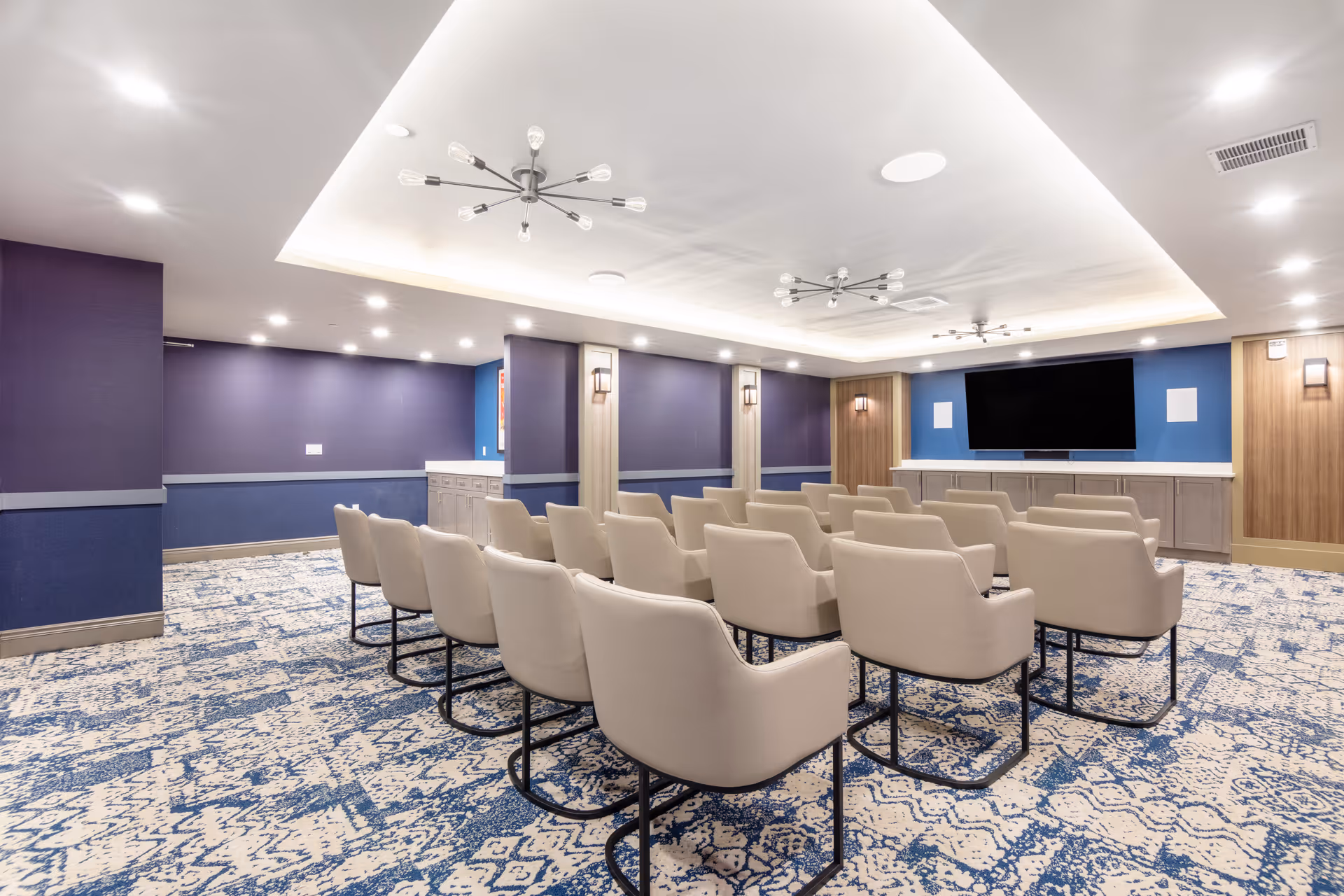 A modern meeting or presentation room with rows of beige cushioned chairs facing a large flat-screen TV mounted on a blue accent wall. The room has patterned blue and white carpeting, recessed lighting, and contemporary ceiling light fixtures. The walls are painted in shades of blue and purple with wooden panel accents.
