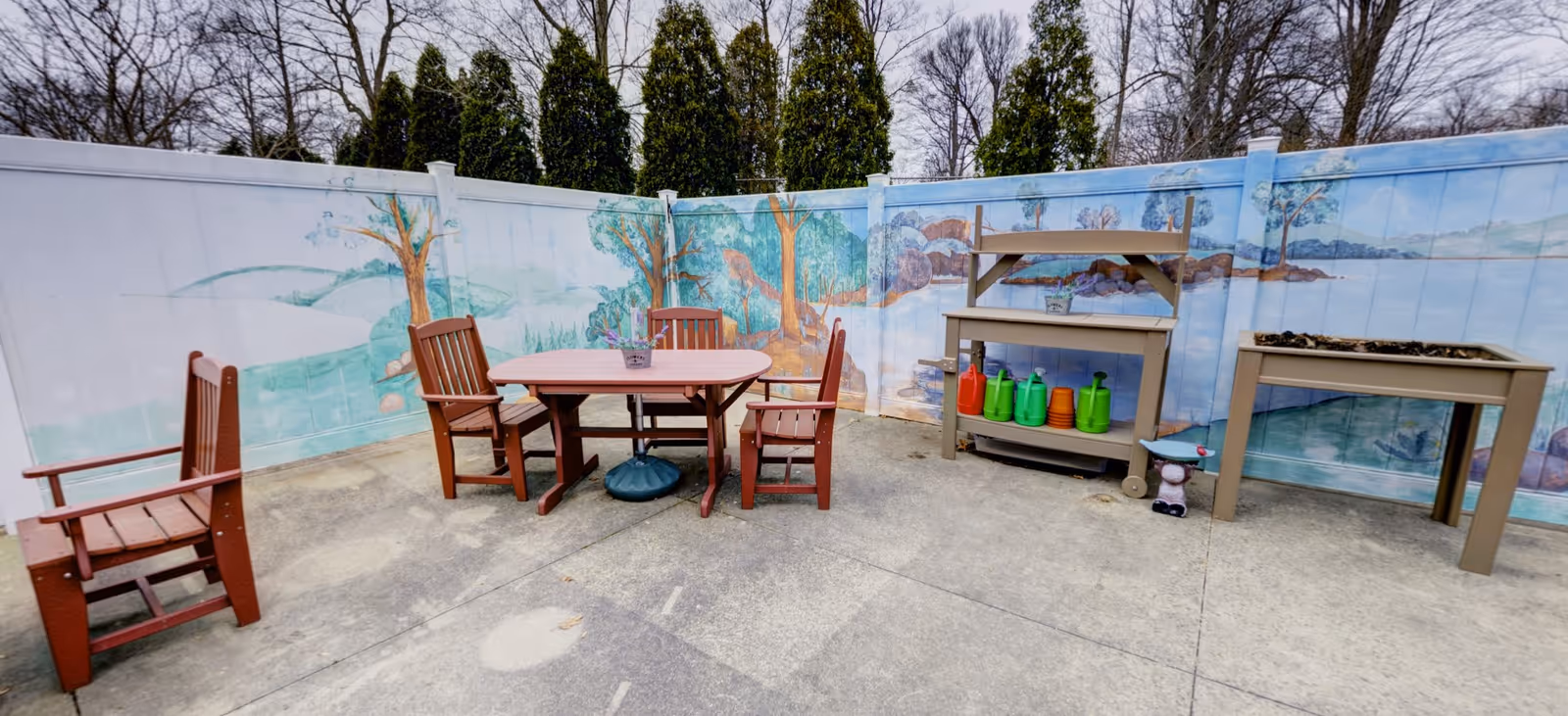 Outdoor patio area with a wooden table and four chairs on a concrete surface. There is a mural painted on the surrounding fence depicting trees, hills, and a river. On the right side, there is a potting bench with colorful watering cans and a planter box. Tall evergreen trees and leafless deciduous trees are visible beyond the fence.