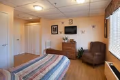 A cozy bedroom with a bed covered in a striped blanket, a wooden dresser with a TV mounted above it, a brown armchair, a white chair, and framed pictures on the wall. The room has wood flooring, a window with blinds, and a ceiling light.