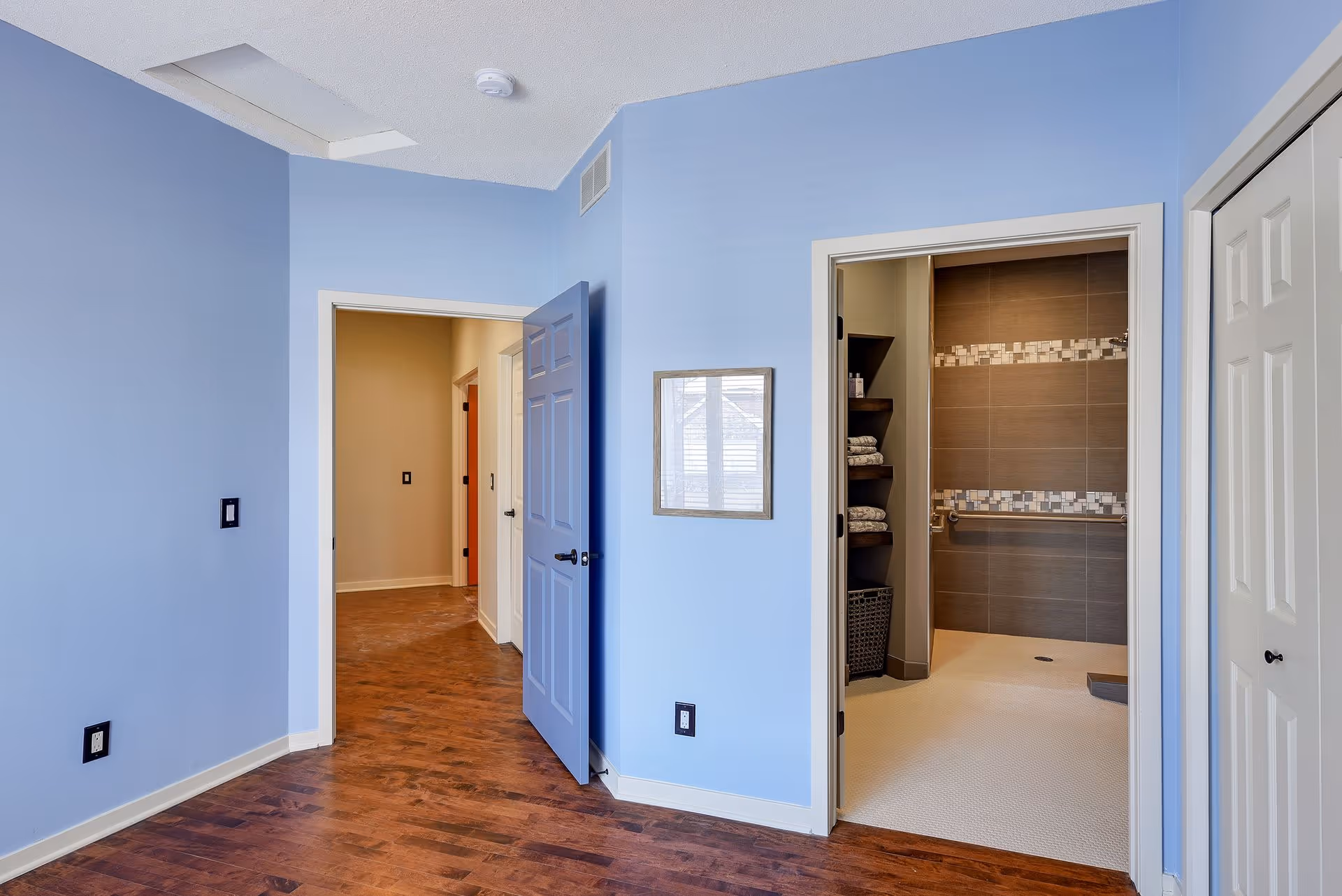 Empty room with light blue walls and wood floors, an open door to a hallway, and an en-suite tiled bathroom with shelving.