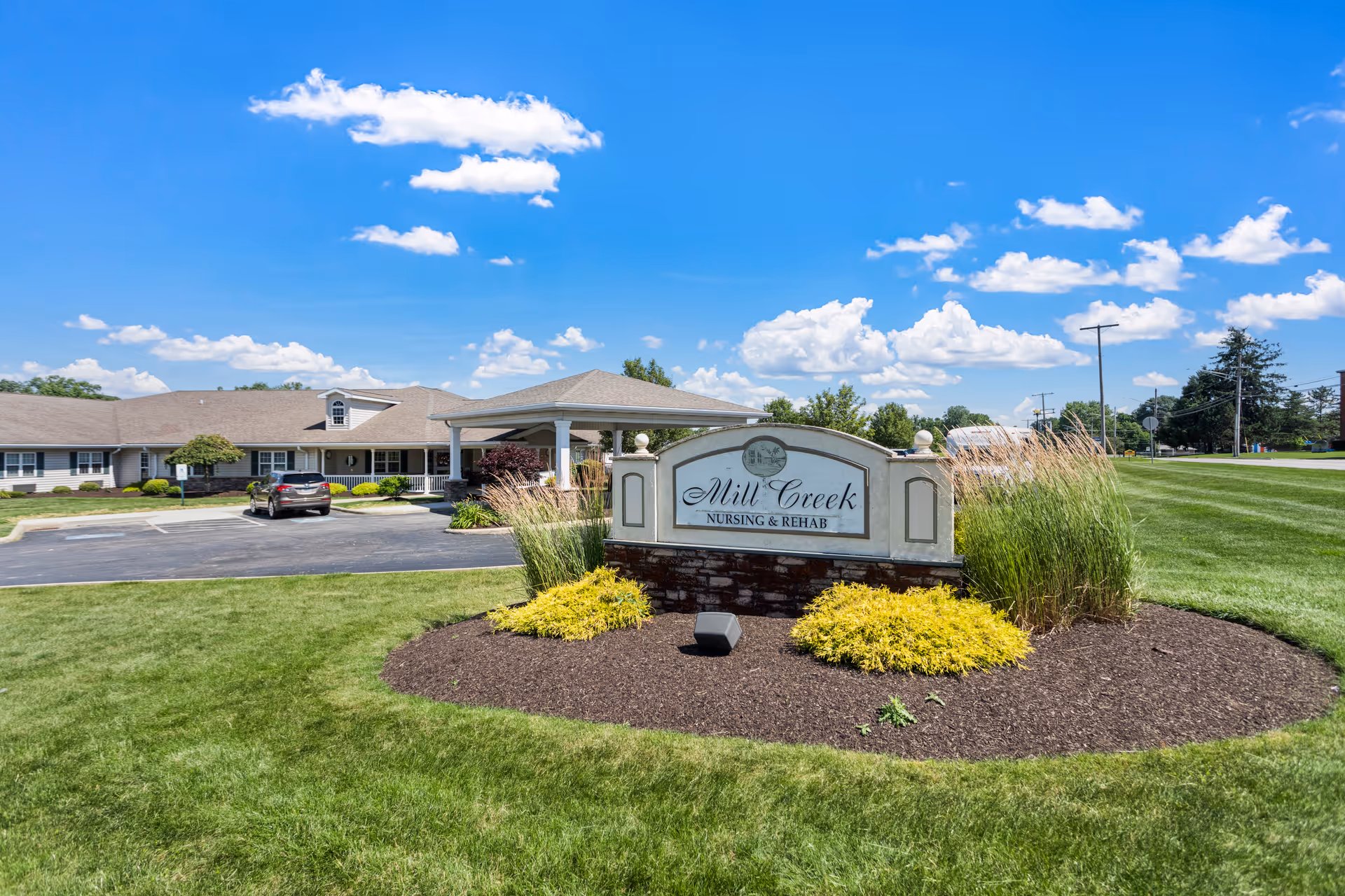 Exterior view of Mill Creek Nursing & Rehab facility on a sunny day with a clear blue sky and scattered clouds. The building is single-story with a covered entrance and a parking lot with a few cars. In the foreground, there is a landscaped area with green grass, ornamental grasses, and yellow shrubs surrounding a stone sign displaying the facility's name.
