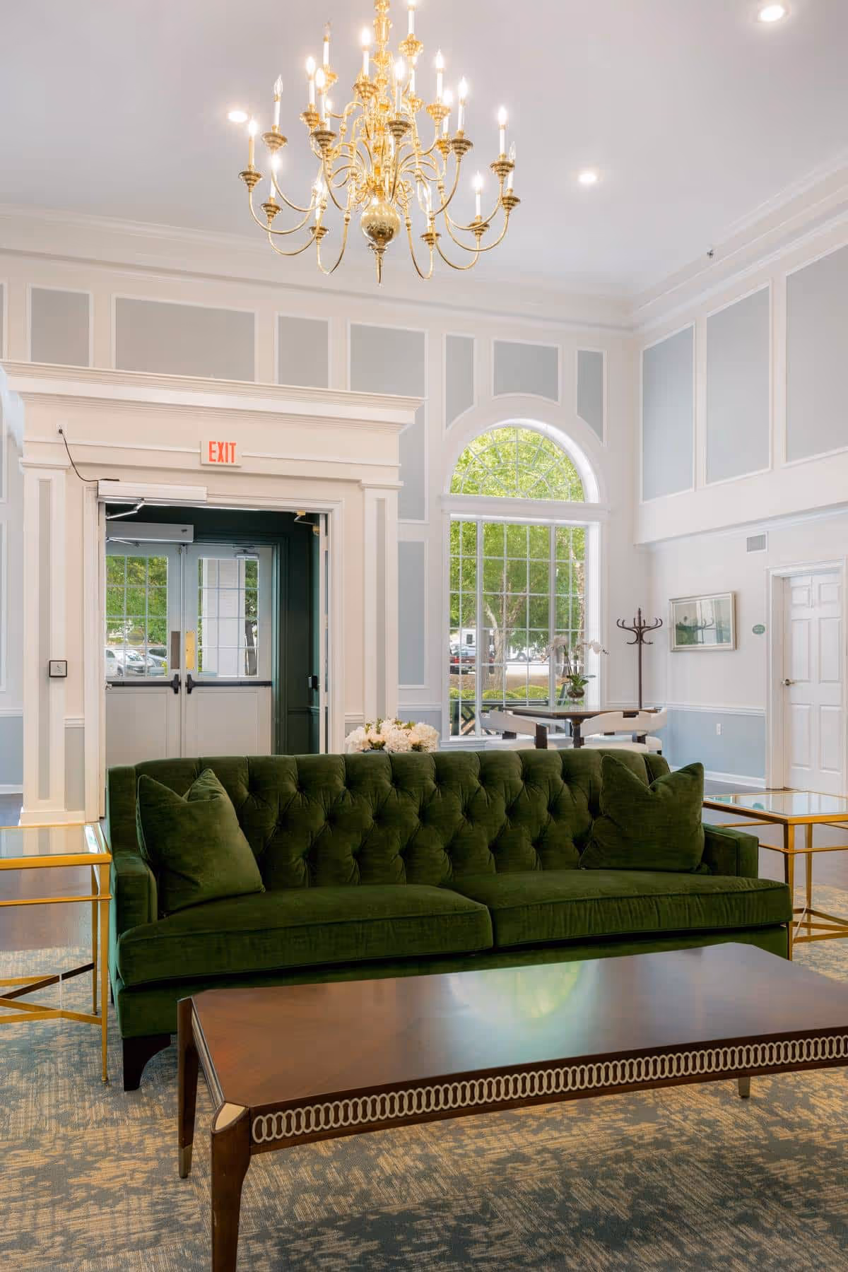 A bright and elegant living room area in Cumberland Hills Assisted Living featuring a green velvet tufted sofa with matching pillows, a wooden coffee table with decorative trim, gold-framed side tables, a large window with an arched top letting in natural light, a chandelier hanging from the ceiling, and light blue and white paneled walls.