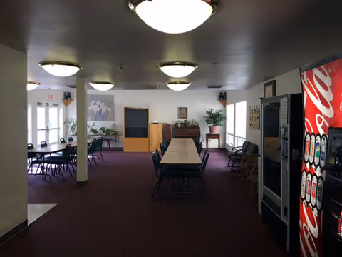 Interior view of a common area in a senior living facility with tables and chairs arranged for group seating. The room has carpeted floors, ceiling lights, vending machines on the right side, and large windows letting in natural light. There are plants and framed artwork on the walls.