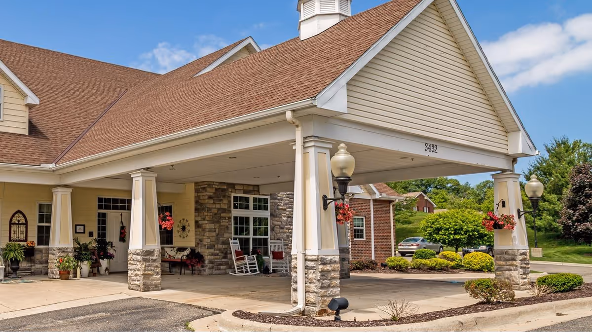 Front entrance of a senior living facility with a covered drop-off area supported by stone and white pillars. The building has a tan exterior with a brown shingled roof. There are hanging flower baskets on the pillars and rocking chairs near the entrance. The address number 3492 is visible on the front of the covered area. Trees and shrubs surround the driveway and parking area under a partly cloudy blue sky.