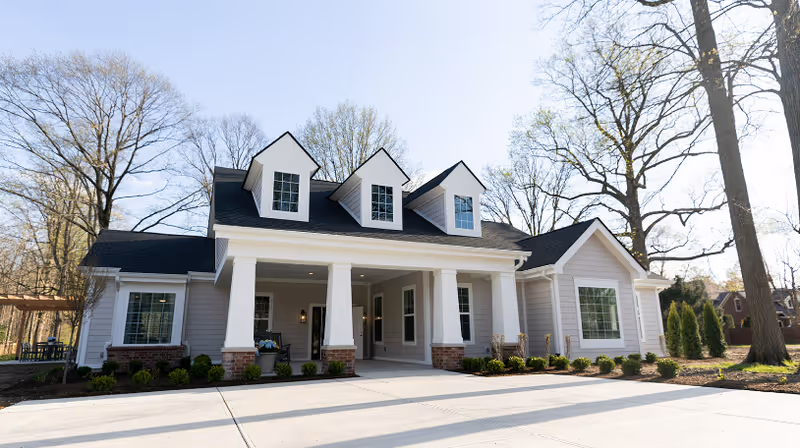 Front exterior of a single-story cottage-style senior living building with white columns, dormer windows, and a paved driveway surrounded by trees.