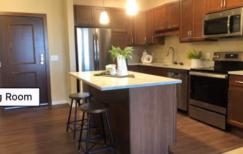 Modern kitchen with wooden cabinets, stainless steel refrigerator, oven, microwave, and dishwasher. A kitchen island with a granite countertop has two black stools and decorative plants on top. The floor is wooden, and there is a door visible in the background.