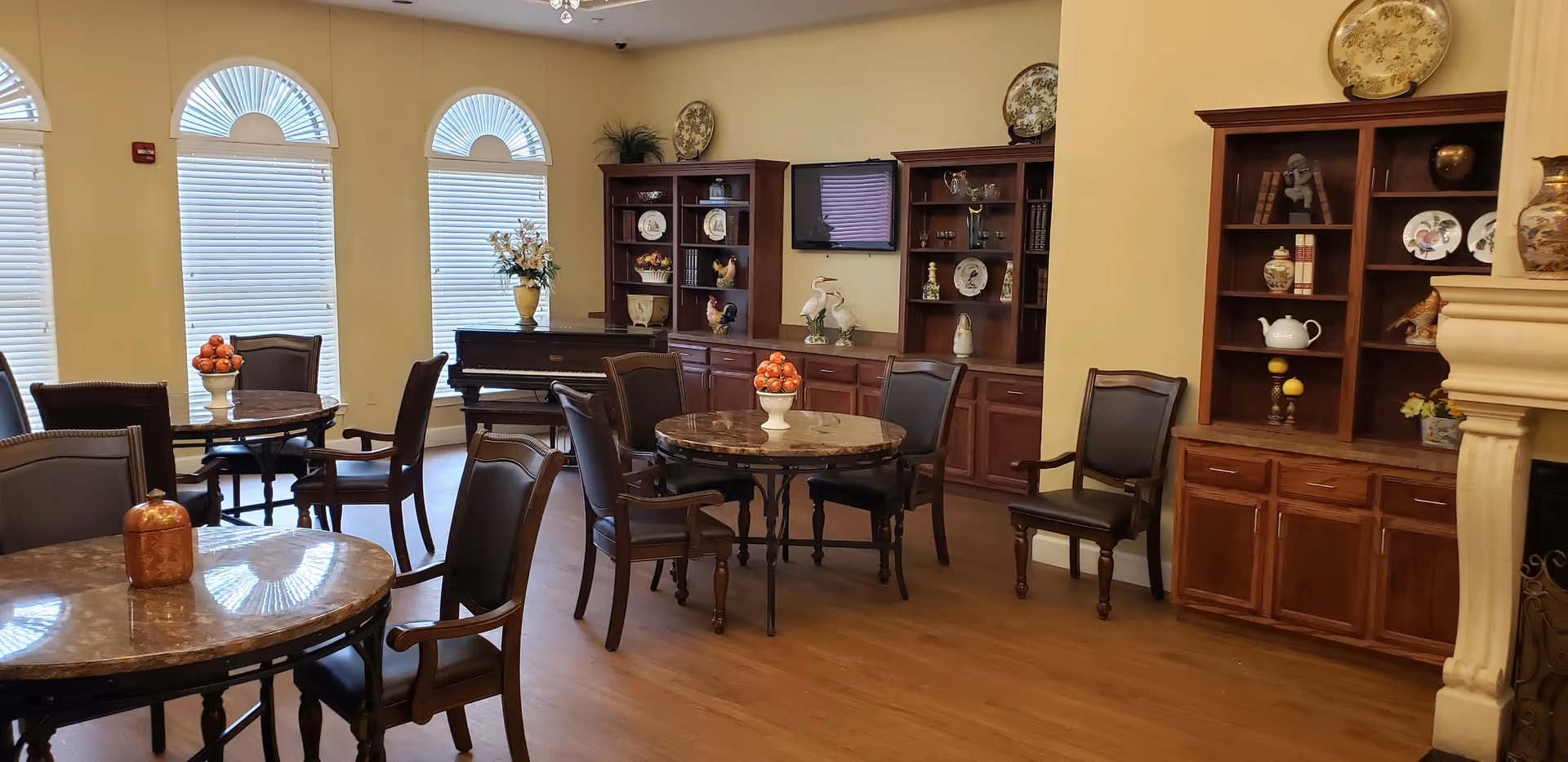 A well-lit common area with round tables and chairs arranged for seating. The room features large arched windows with blinds, wooden cabinets and shelves displaying decorative plates and vases, a piano, and a wall-mounted TV. The floor is wooden, and the walls are painted a soft yellow.