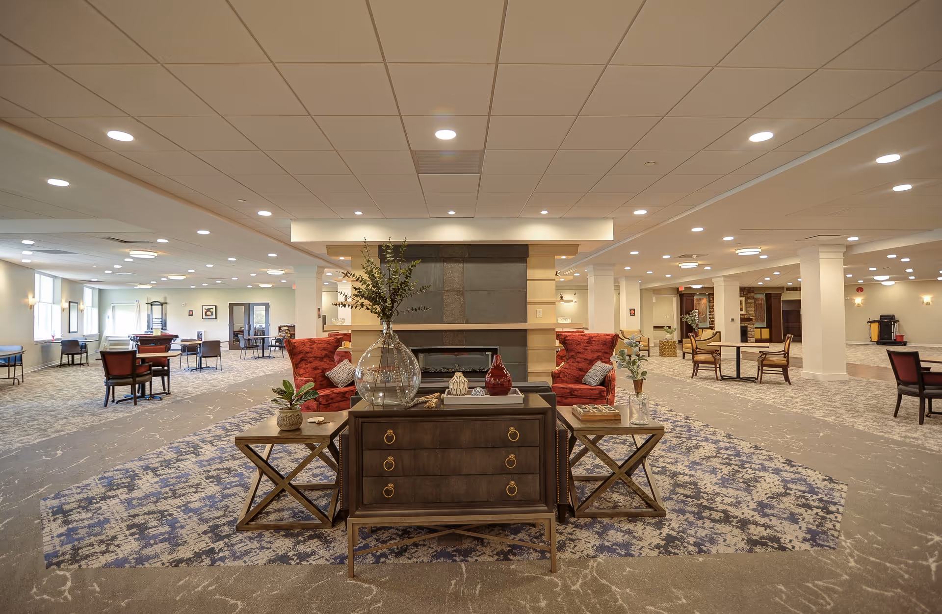 Spacious and well-lit common area in a senior living facility featuring a central fireplace with decorative vases on a wooden console table, surrounded by red upholstered armchairs. The room has multiple seating areas with tables and chairs, carpeted floors with patterned rugs, and recessed ceiling lights.