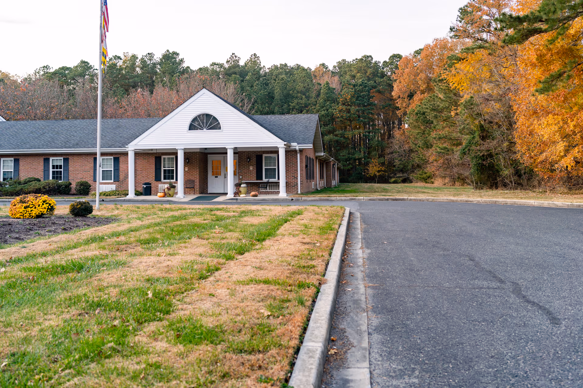 Front exterior view of a single-story brick building with white columns at the entrance, a flagpole with flags, and autumn trees in the background.