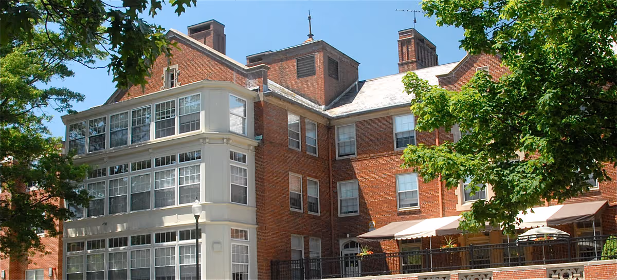 Exterior view of a large brick building with multiple windows and a white-framed sunroom or enclosed porch area. The building is surrounded by green trees under a clear blue sky.