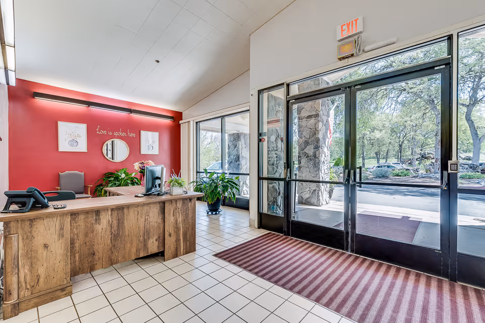 Reception area of Meadowood Nursing Center with a wooden desk, computer, phone, and plants. Behind the desk is a red wall with two framed pictures, a round mirror, and the phrase 'Love is spoken here'. To the right are glass double doors leading outside, with a striped red and brown floor mat in front of them.