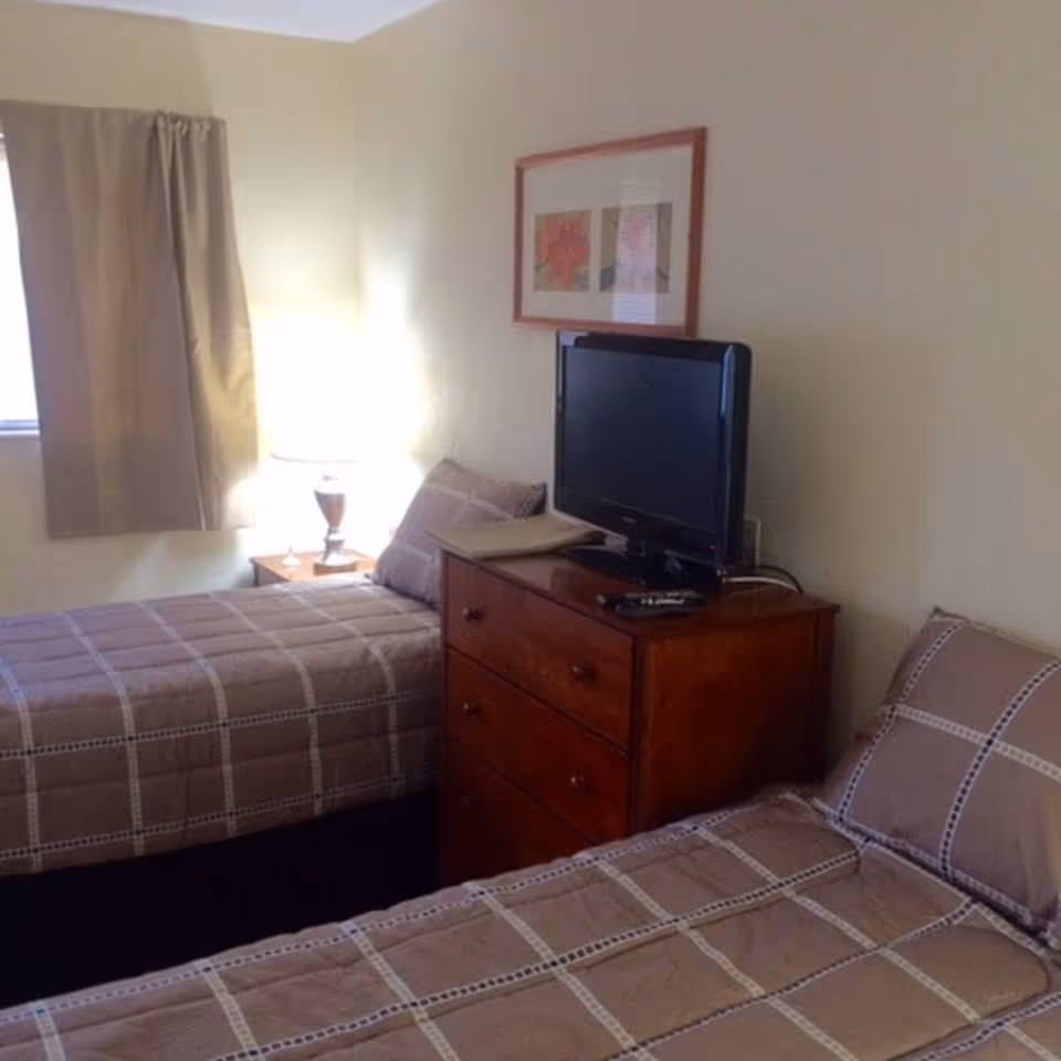 A bedroom with two single beds covered in matching brown and beige checkered bedding. Between the beds is a wooden dresser with a flat-screen TV on top. A lamp and framed artwork are visible on the wall behind the dresser. A window with beige curtains is on the left side of the room.