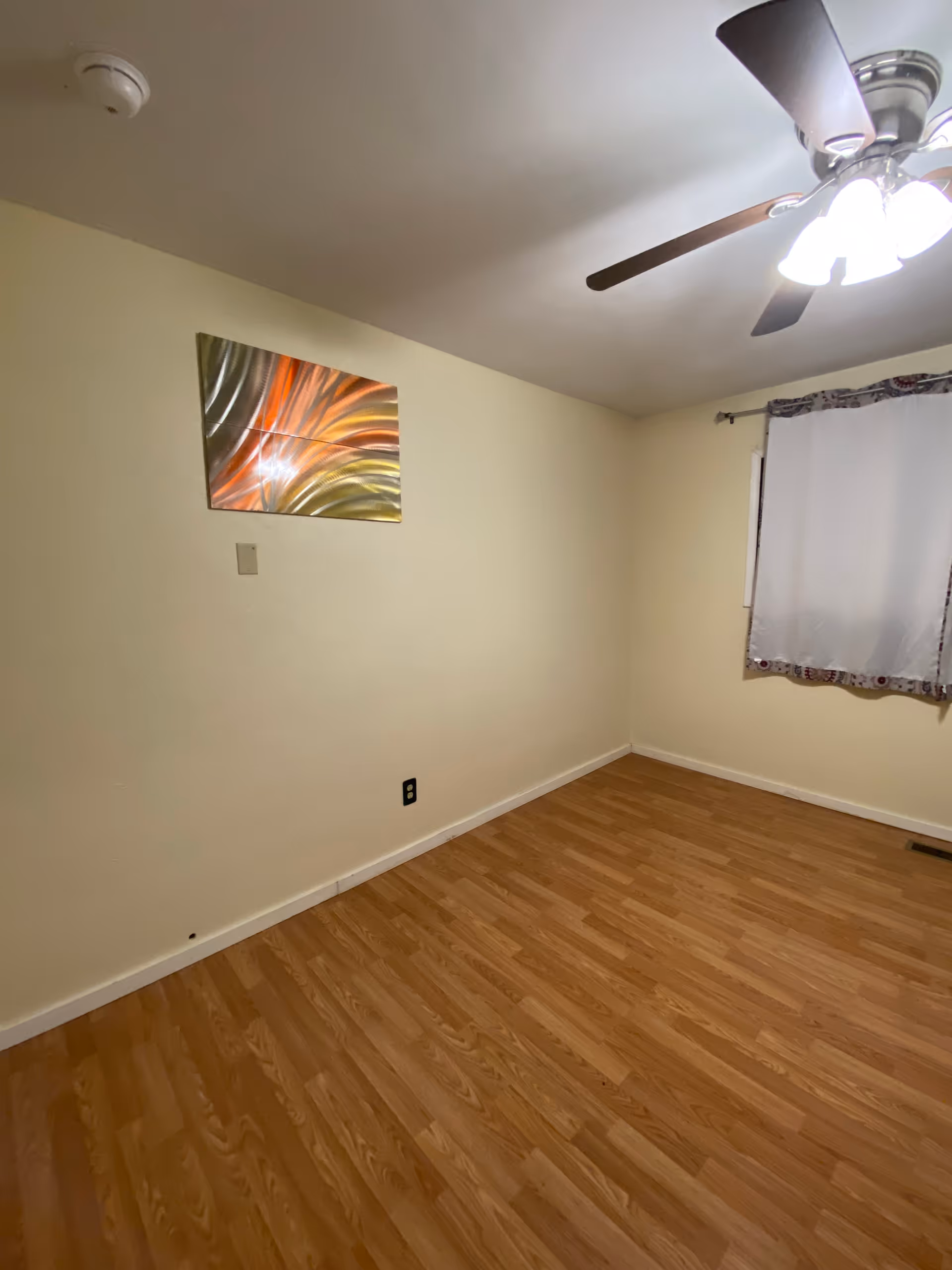 Empty room with light wood laminate flooring, cream-colored walls, a ceiling fan with lights, a window with a white curtain, and a colorful abstract metal wall art piece.