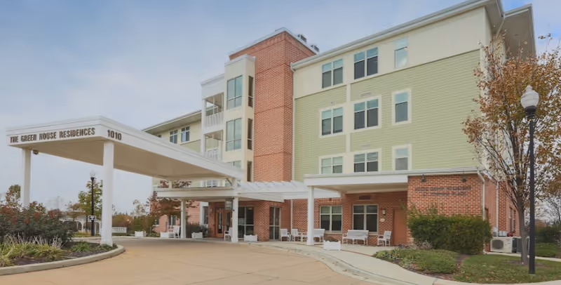 Exterior view of a multi-story residential building with a covered entrance labeled 'The Green House Residences 1010'. The building features a combination of brick and light green siding, with several windows and outdoor seating areas. There are trees and landscaped greenery around the driveway and entrance.