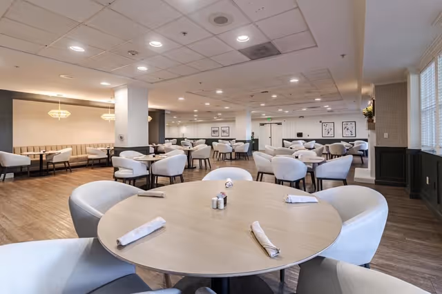 A spacious dining room with multiple round tables and cushioned chairs arranged neatly. Each table has rolled napkins and salt and pepper shakers. The room features wood flooring, soft lighting from ceiling fixtures, and framed artwork on the walls. Large windows on the right side allow natural light to enter the space.