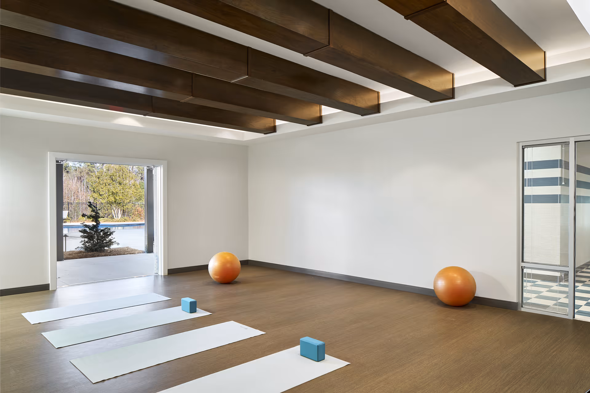 A spacious exercise room with wooden flooring and white walls. The ceiling features large dark wooden beams. There are three white yoga mats laid out on the floor, each with a blue yoga block. Two large orange exercise balls are placed against the walls. A large open doorway leads outside to a pool area with trees visible in the background. A glass door with blinds is on the right side of the room.