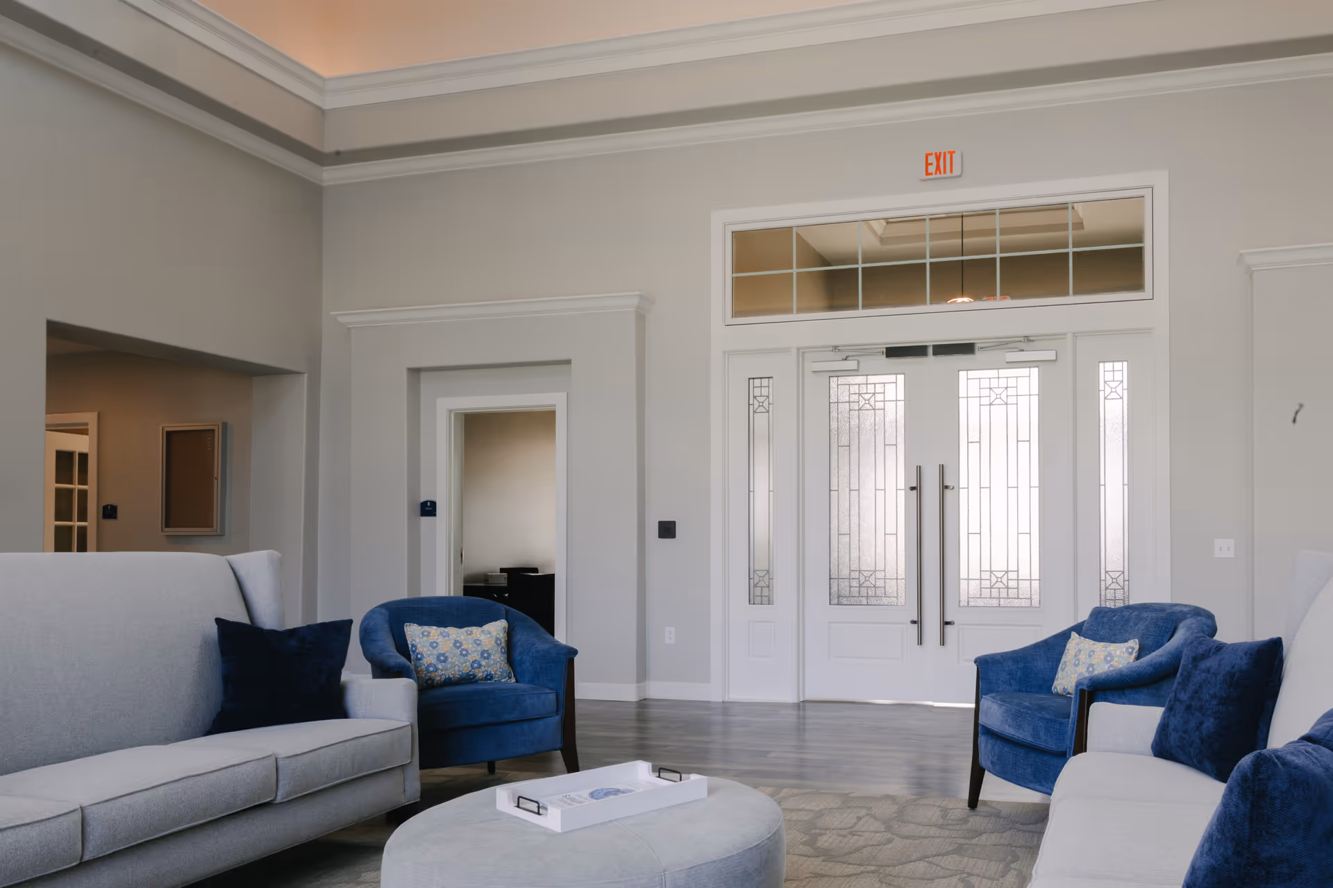 A bright and spacious living room area with light gray walls and high ceilings. The room features two gray sofas and two blue armchairs with patterned cushions arranged around a round gray ottoman with a white tray on top. In the background, there is a set of double doors with decorative glass panels and an exit sign above them. The floor is covered with a light-colored rug and wood-like flooring.