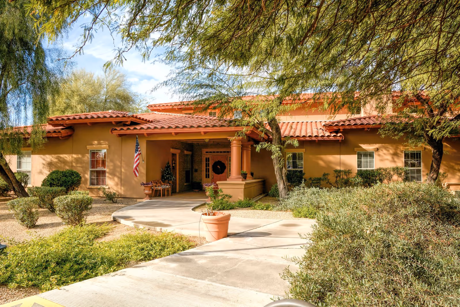 Exterior view of Barton House facility with a tan stucco building featuring a red tile roof, surrounded by trees and shrubs. A concrete walkway leads to the entrance, which has a covered porch with columns, an American flag, and some outdoor seating.