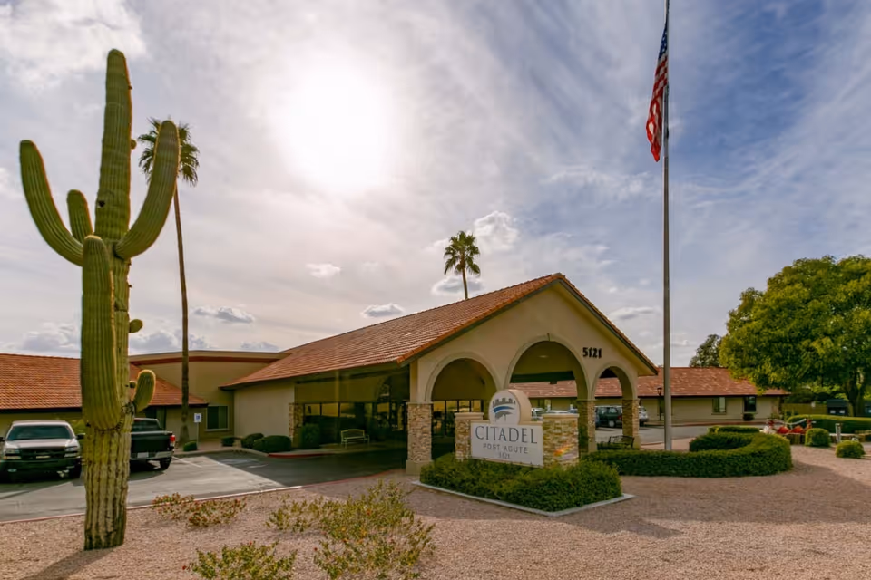Exterior view of Citadel Post Acute facility with a large cactus and palm trees in the foreground, a flagpole with an American flag, and a clear sky with some clouds.