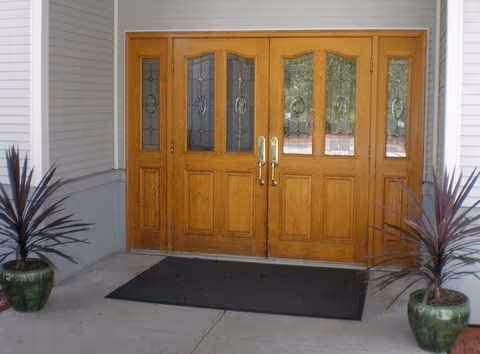 Double wooden entrance doors with glass panels flanked by potted plants and a doormat.