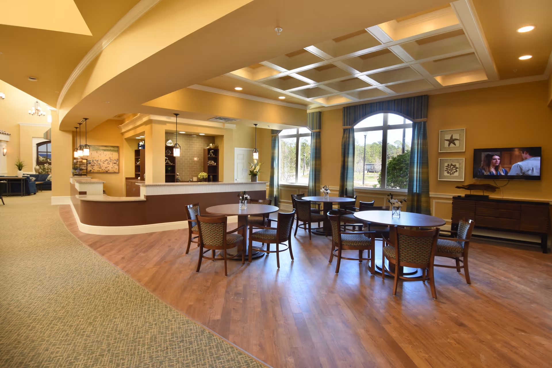 A spacious dining area in a senior living facility with round wooden tables and chairs arranged on a wooden floor. Large windows with blue curtains allow natural light to fill the room. The ceiling features a decorative coffered design with recessed lighting. A television is mounted on the wall above a wooden cabinet, and a counter area with pendant lights is visible in the background.