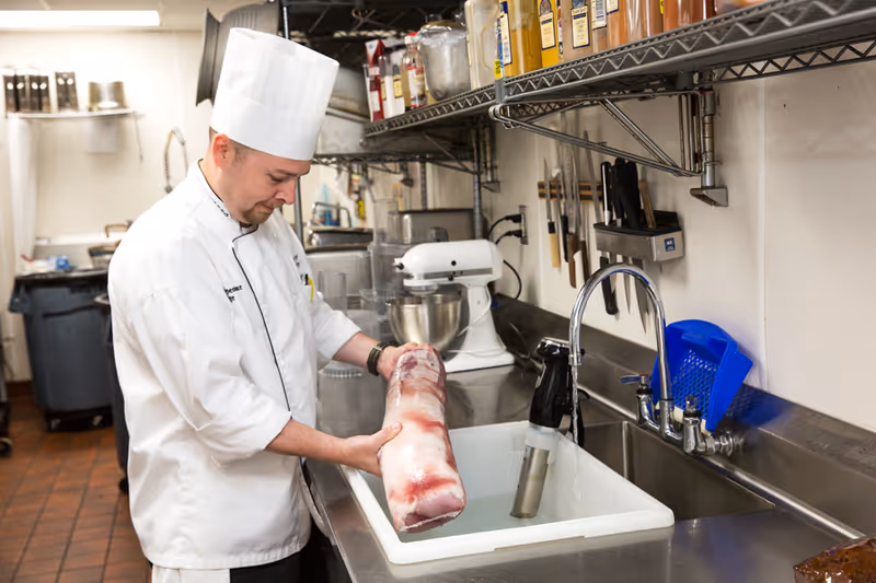 A chef wearing a white uniform and tall white chef hat is holding a large piece of meat over a sink in a commercial kitchen. The kitchen has metal shelves with various spices and utensils, a mixer, and knives hanging on the wall.
