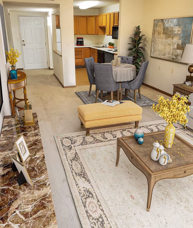 Interior view of a senior living facility showing a living room and dining area. The living room has a beige carpet, a wooden coffee table with decorative items including a yellow vase with flowers, and a yellow upholstered bench. The dining area features a round table with a tablecloth and four gray upholstered chairs on a small rug. The kitchen with wooden cabinets and appliances is visible in the background. The walls are decorated with a large abstract painting and a tall green plant. A small wooden side table with a blue vase and yellow flowers is near the entrance door.