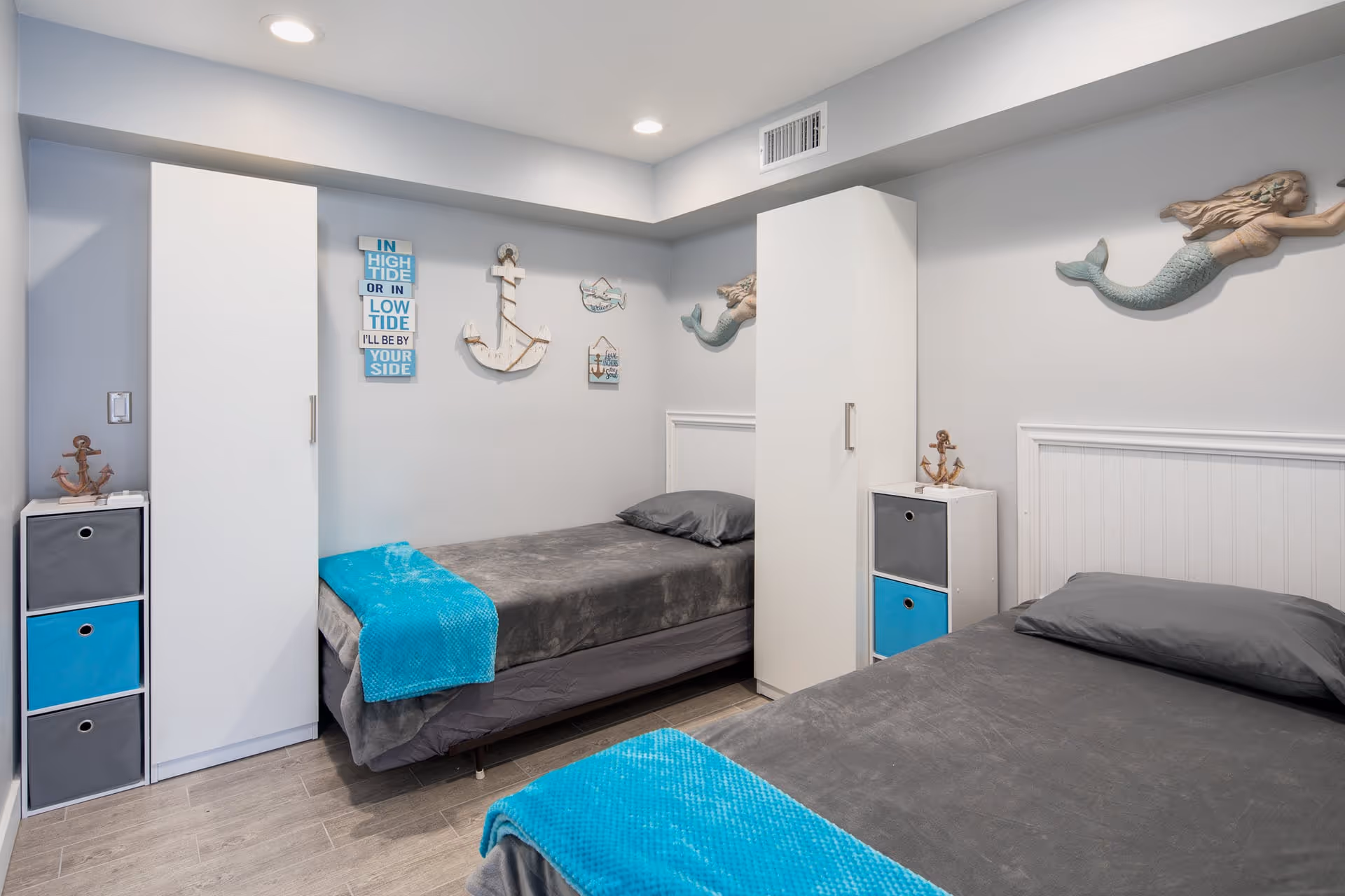 A small bedroom with two single beds, each covered with gray bedding and a bright turquoise throw blanket. The room has light gray walls decorated with nautical-themed wall art including anchors, a mermaid, and a sign that reads 'IN HIGH TIDE OR IN LOW TIDE I'LL BE BY YOUR SIDE'. There are two white wardrobes and small storage units with gray and blue drawers beside each bed. The floor is light wood, and the ceiling has recessed lighting.